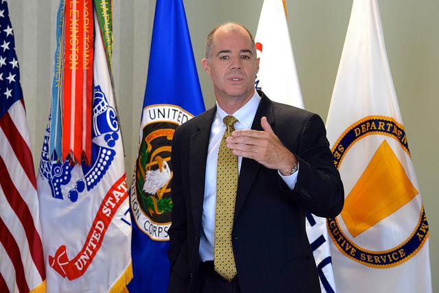 Man gestures while speaking in front of flags.